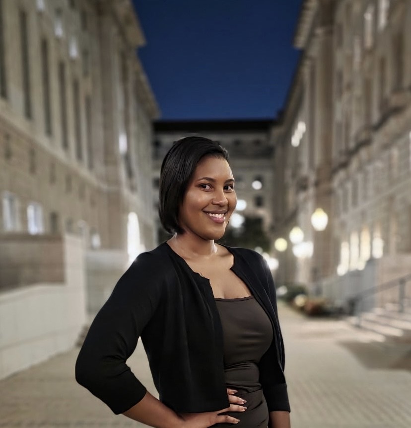 Professional photo of editor Cecilia Gray wearing a gray tank top and black long-sleeve cover with her hand on her hip. She is standing in front of a GWU building.