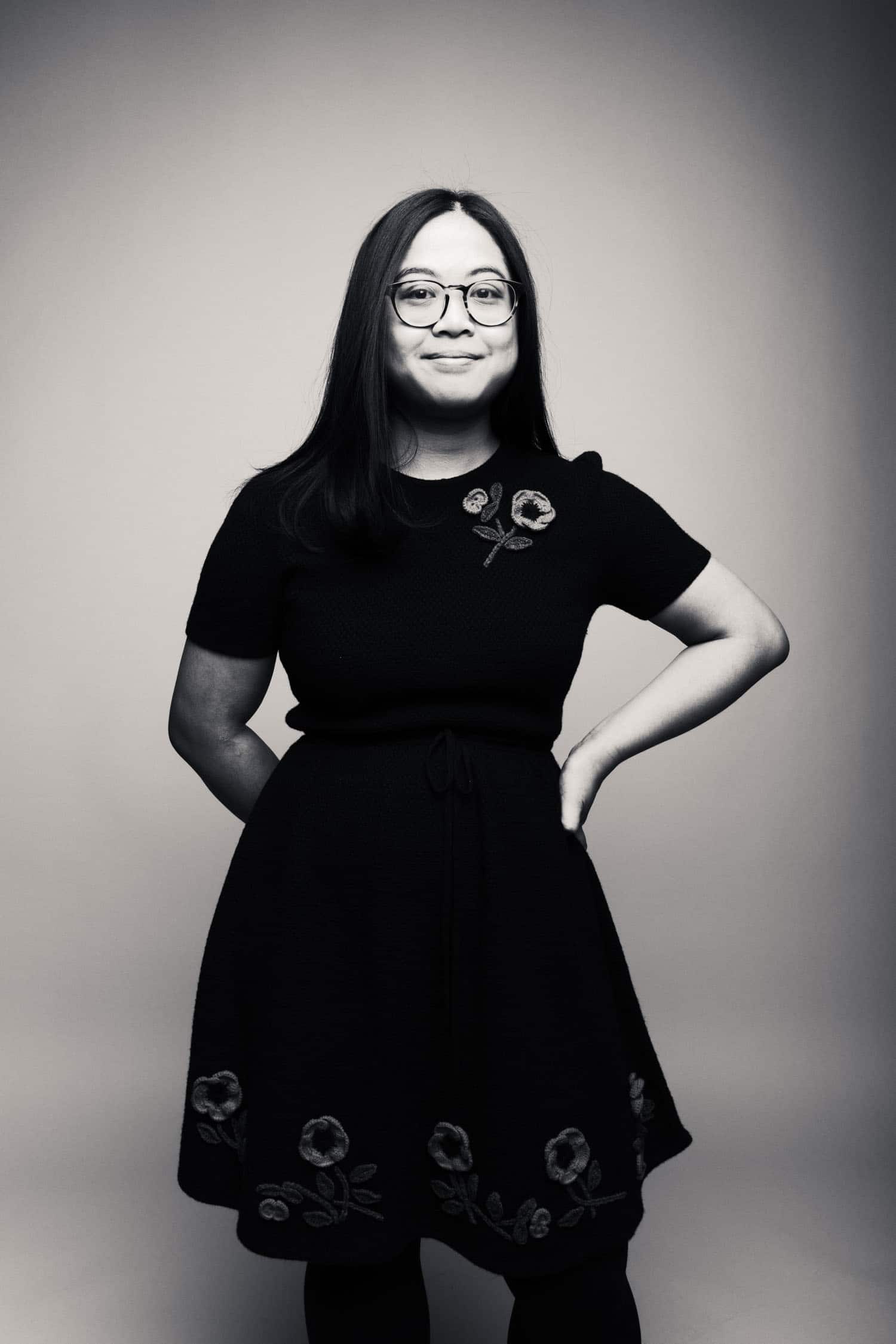 Author Kate Gavino wearing a black dress with flowers along the bottom, posing with hands on hips in this black and white photo.